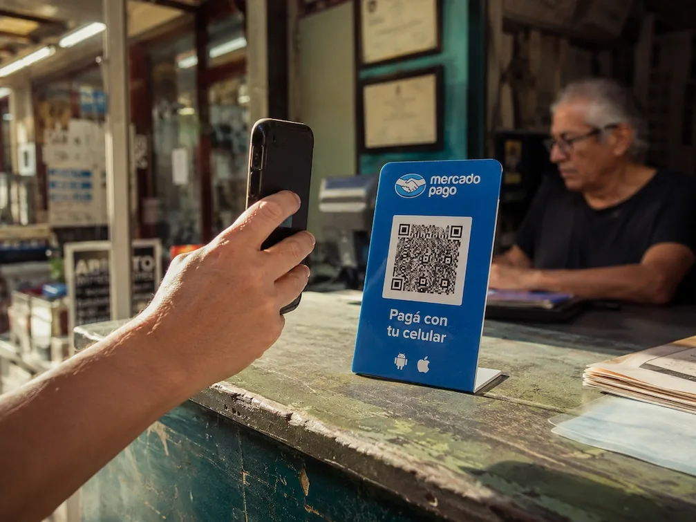 A hand holds a phone up to the blue Mercado Pago QR standee on a Buenos Aires shop counter.
