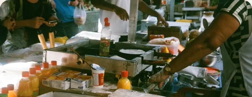 A vendor's hands work a hot griddle at a busy Brazilian street food market stall.